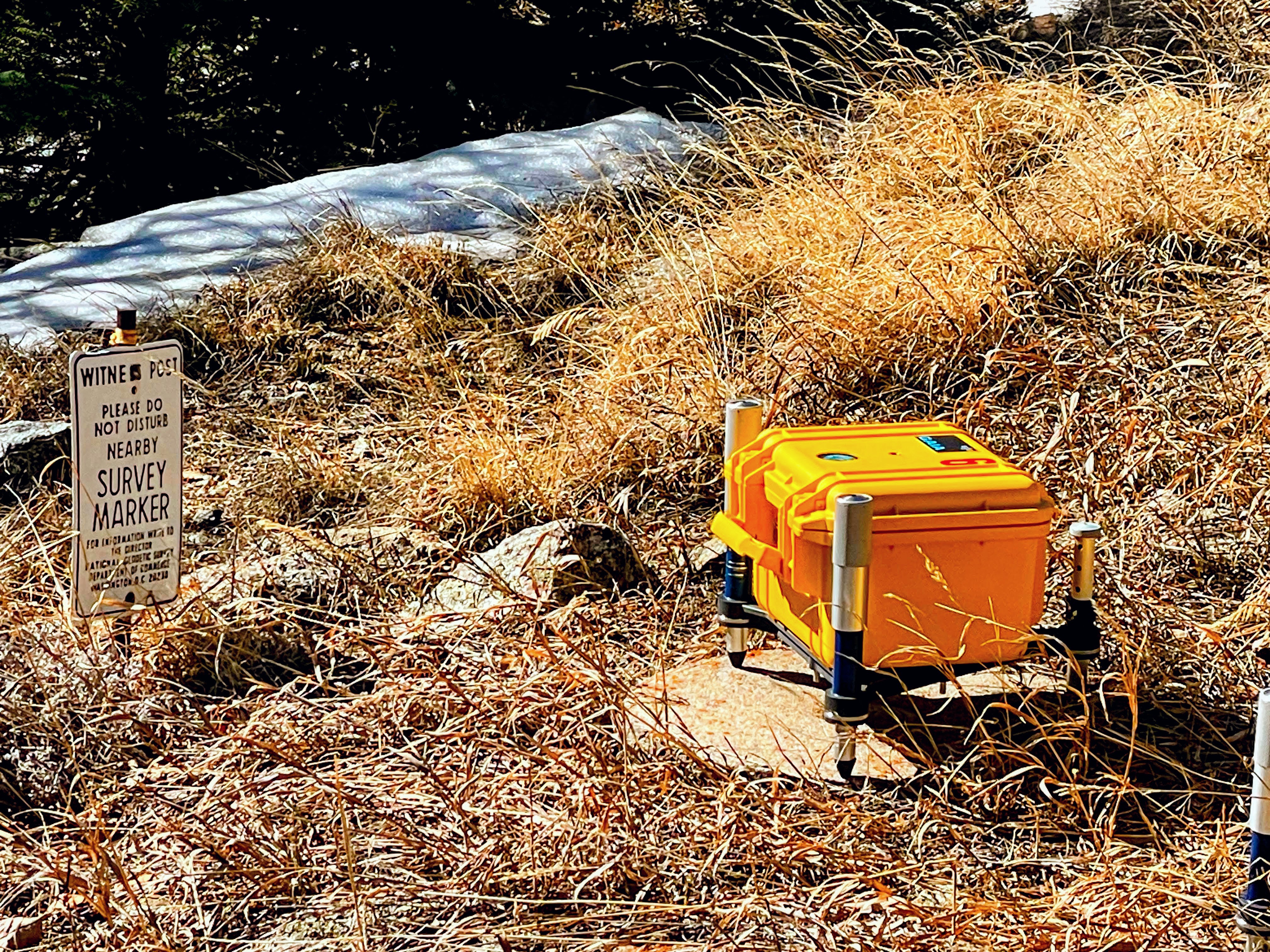 A yellow survey instrument is set up on tripod legs amid dry grass near a sign indicating a survey marker. Patches of snow are visible in the background.